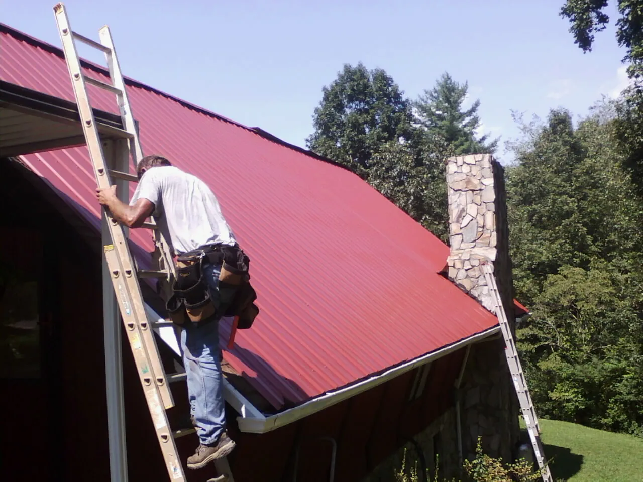 Roofer on a ladder beside a red metal roof and stone chimney — Soffit & Fascia Repair by Roofing Brothers in Bush